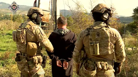 SSG Two soldiers detain a man in handcuffs wearing dark clothes in a lay-by.