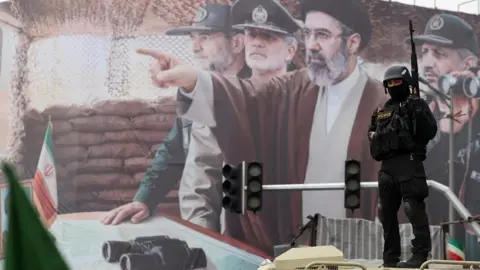 EPA A member of the Iranian security forces stands guard in front of a billboard depicting the new Supreme Leader, Mojtaba Khamenei, and Iranian military and Revolutionary Guards commanders, during a rally to mark Quds Day at a square in Tehran, Iran (13 March 2026)