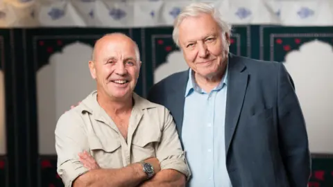 PA Media Doug Allan, left, wearing a beige shirt, with his arms folded across his chest. He is bald and is smiling at the camera. He is wearing a silver watch on his right wrist. Sir David Attenborough, right, wearing a grey suit jacket over a blue shirt. He is smiling and has white hair. He is standing in front of a white and green background.