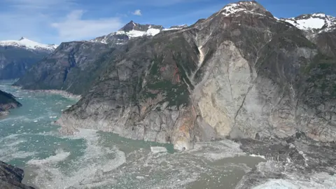 Photo by Cyrus Read/U.S. Geological Survey An icy expanse of sea runs through a narrow passage between mountains. Much of the steep sides of the mountains are stripped of green vegetation revealing grey scarred rock. The tops of the mountains are scattered with snow and ice glitters in the fjord below. 