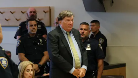 James Carbone/Pool via REUTERS Gilgo Serial Killer Rex Heuermann stands in a dark jack, white shirt and yellow tie. He is speaking in a courtroom, and there are several police in uniform around him. A woman is sitting at a table next to him.