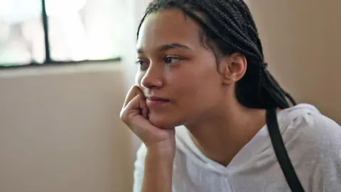 Cathy has dark hair tied back in braids and is wearing a white top with beige walls and a window in the background