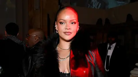 Getty Images Rihanna has short hair wearing a black furry coast with a red light cast on her face