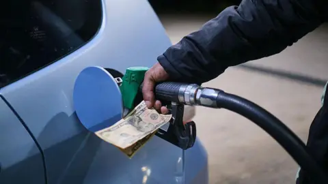 AFP via Getty Images A hand holding a petrol pump into a car as it fills up at a petrol station in Zimbawe. The same hand also holds paper money. The car is silver.