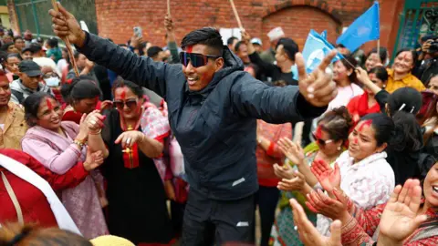 Getty Images Rastriya Swatantra Party supporters celebrate following one of their candidates' win. A young man wearing sunglasses and a dark jacket and trousers jumps up and down with joy as he is surrounded by a crowd clapping and chanting