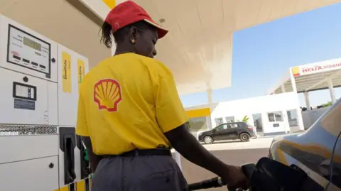 AFP via Getty Images The back of a woman wearing a yellow T-shirt with a Shell logo on the back and a red cap stands by a car as she fills it up with petrol.