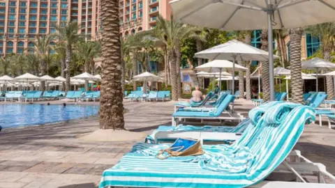 Getty Images Blue-striped sun loungers covered in towels are lined up alongside a turquoise hotel swimming pool.