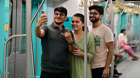 Hindustan Times via Getty Images Two young men and a woman who is between them, take a selfie while traveling on the Aqua Line metro train in Mumbai. 