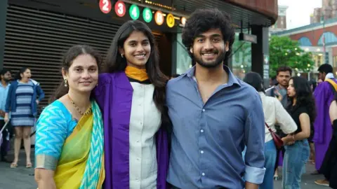 Bhavitha Mandava/Instagram Bhavitha Mandava with her mum - who's dressed in a yellow/blue sari - and brother - wearing a blue shirt - as she poses on a busy street 