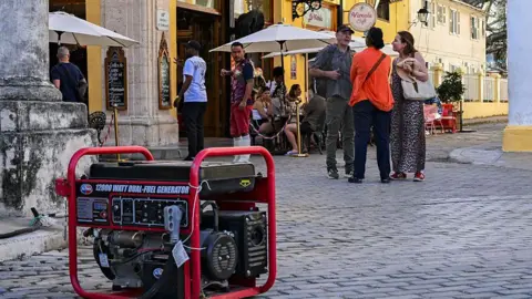 Getty Images A shopkeeper uses a generator during a blackout. The generator is on cobblestones and there is a group of people nearby