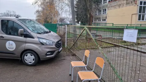 De Akcióközösség A van stands outside a school gate in a Hungarian village, with two chairs on the ground