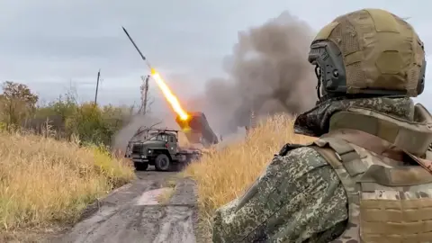 EPA A road with overgrown, dry grass on either side. At the end of the road (in the centre back) is a military vehicle and a rocket being launched. In the front right, is the back of a soldier watching it. The soldier is wearing military fatigues with a vest and a helmet.