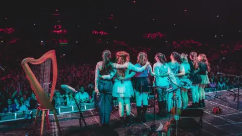 Claire Loughran Music group - eight women stand arm in arm on a concert stage facing a large stadium audience, with instruments including a harp visible at the side under colourful stage lighting.
