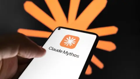 Getty Images A person's hand holding a smartphone using Anthropic's Claude Mythos app. The words 'Claude Mythos' appear prominently on the white screen with the company logo (a white, cartoonish asterisk on an orange square with curved edges) above. Behind the hand and the phone in soft focus is another version of the logo (an orange, cartoonish asterisk) on a black wall.