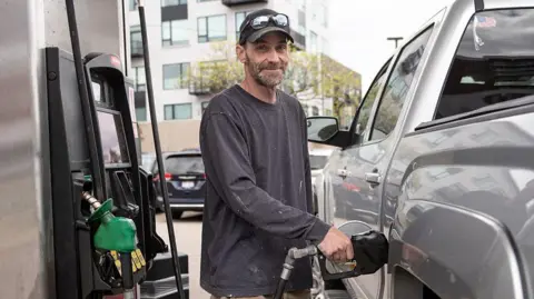 Adam J. Dewey/Anadolu via Getty Images A man smiles for the camera as he fills up his grey pick-up truck at a petrol station 