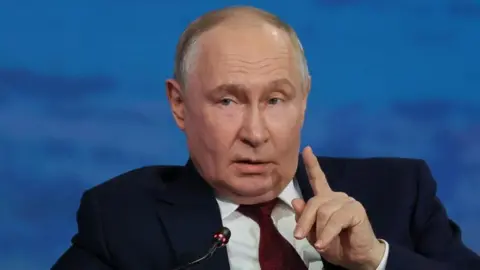 Reuters President Putin lifts his finger as if to make a point as he speaks into a microphone. He is dressed in a navy blue suit, white shirt and red patterned tie and is speaking in front of a marine blue backdrop