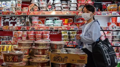 AFP via Getty Images A customer walking past a row of shelves filled to the brim with instant noodles at a supermarket in Japan.