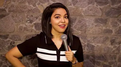 Ankita Shrivastav Ankita Shrivastav, wearing a black-and-white T-shirt, is standing on a stage and holding a mic in her hand.