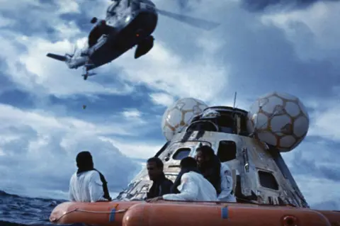 Getty Images Apollo 13 astronauts waiting for rescue in a raft. The space capsule is shown behind them and a helicopter is hovering overhead.