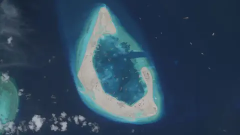 Getty Images Vantor satellite imagery shows an overview of Antelope Reef, including a partially enclosed lagoon, reclaimed sand areas, and numerous vessels operating around the feature. The image provides broader context of ongoing visible expansion and marine activity at the reef.