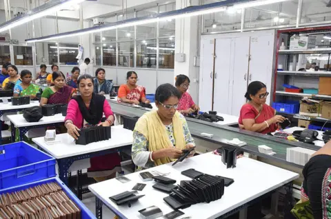 NurPhoto via Getty Images Women wearing colourful Indian clothes sit at white tables. Each of them has piles of black leather wallets in front of them