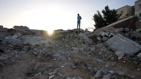 Neha Sharma/BBC A man stands on top of rubble from a building destroyed in an Israeli air strike in Saksakiyeh, southern Lebanon, which killed nine members of one family