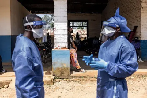 Getty Images The image shows a partially open building with a shaded interior and a dirt ground outside. In the foreground, two individuals are standing and wearing full protective gear, including blue gowns, gloves, face masks, and clear face shields. One individual appears to be gesturing with gloved hands, while the other faces toward them, suggesting a conversation or coordination. In the background, several people are sitting or standing under the roofed area, some near parked motorcycles and a wall with a window. 