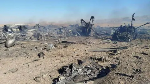 Reuters A still image purporting to show the burned wreckage of aircraft destroyed during the US mission to find a stranded airman in Iran. What appears to be two propellers can be seen among scraps of metal and smoke on a sandy ground.