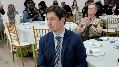 BBC / Brian Khisa Minneapolis Mayor Jacob Frey sitting at one of several round dinning tables with white table clothes in a room hosting a community event. Among those seated behind him can be seen state security officers.