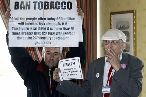 Getty Images David Hockney and an anti-smoking protester in 2005. The protester is holding up a sign calling for tobacco to be banned. Hockney is holding up a small counter sign saying that people die even if they don't smoke. He is smoking.