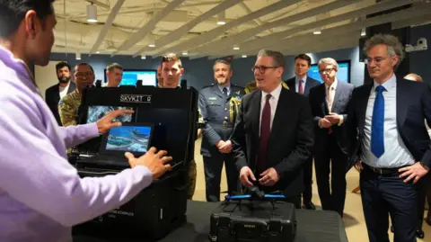 Getty Images UK Prime Minister Sir Keir Starmer stands next to Palantir boss Alex Karp during a Washington DC visit in February 2025 while being shown a demo of Palantir "Skykit" tech. Peter Mandelson, the UK ambassador at the time, is among a group of men in suits and military uniforms standing behind them.