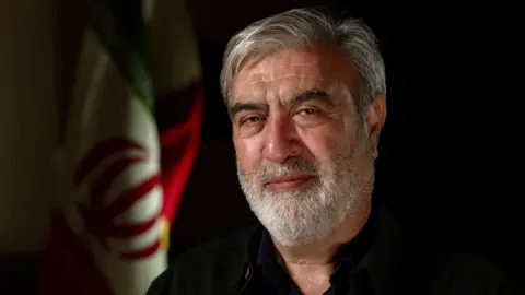 BBC A man with a grey beard and hair looks into the camera. He is wearing a black shirt and behind him is the Iranian flag.