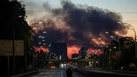 Reuters A man stands on an empty highway to photograph dark clouds of smoke rising through the early morning sky over a city.