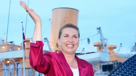 EPA Danish Prime Minister Mette Frederiksen waves, smiling, in a bright pink blazer. She stands in front of the royal yacht Dannebrog. 