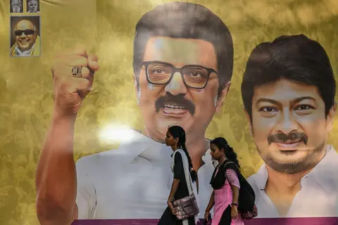AFP via Getty Images Women walk past a poster featuring MK Stalin (C), Chief Minister of Indian state of Tamil Nadu and Deputy Chief Minister Udhayanidhi Stalin along a road in Chennai on March 15, 2026. 