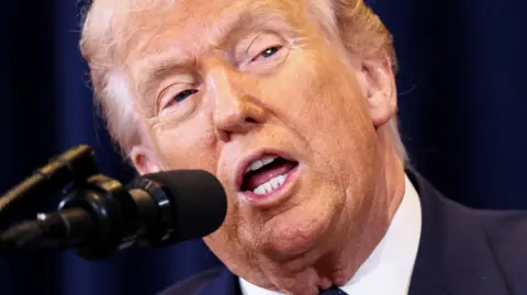 Reuters US President Donald Trump speaks into a microphone, his mouth is agape and he is wearing a blue suit jacket and white shirt. Behind him is a dark blue curtain.