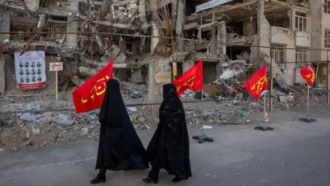 Getty Images Two women in black head-to-toe walk past destroyed buildings and a series of red flag with yellow Arabic script. 