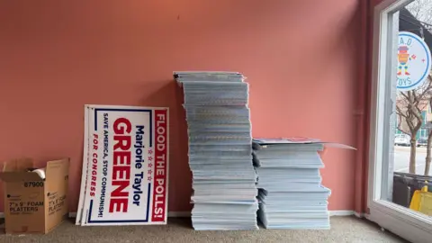 A stack of campaign signs sits on a carpet in an empty office. The signs read: "Flood the polls! Marjorie Taylor Greene: Save America, Stop Communism!"