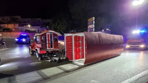 Ayuntamiento Cártama A small red tourist train, with its passenger wagon lying sideways on the street. Some emergency service vehicles can be seen near it.
