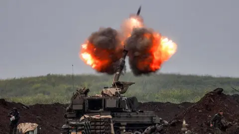 AFP via Getty Images An Israeli self-propelled howitzer artillery gun fires rounds, with balls of fiery smoke around the weapon shooting into the air. The machine is surrounded by a dirt trench, with a grassy hill in front of it.