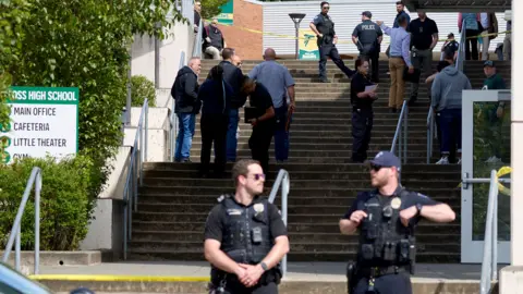 AP Two police officers stand at the bottom of concrete steps outside of Foss High School, facing the camera. A sign reading 'Foss High School' is to their left. Behind them stand around 14 people wearing civilian clothing and more police on the steps, with yellow police tape visible at the top of the steps.