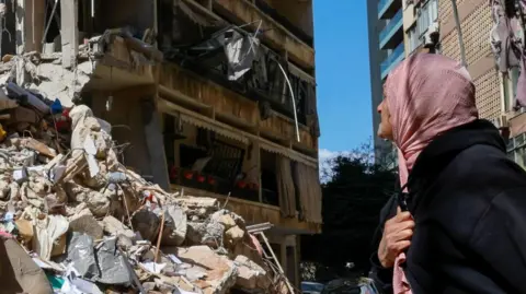 Reuters A woman clutching her head scarf looks at a building destroyed by an Israeli air strike, Beirut (09/04/26)