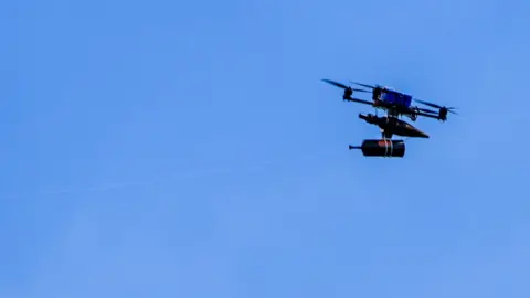 Reuters A fibre-optic drone flies in a clear blue sky across the border from Lebanon into Israel. It has a spool of fibre optic wire hanging under a chassis, supported by four propeller blades around a blue rectangular battery. 