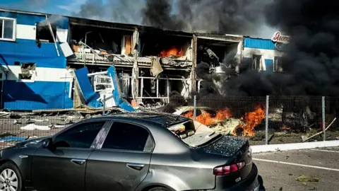 EPA/Shutterstock Black smoke is seen rising from a building, where some of the rooms are exposed and flames can be seen inside. A car is on fire in front and another car is seen in the foreground. 