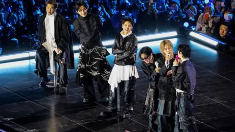 Getty Images RM, seated, while V, Suga, J-Hope Jimin and Jin stand on stage at the comeback show . 