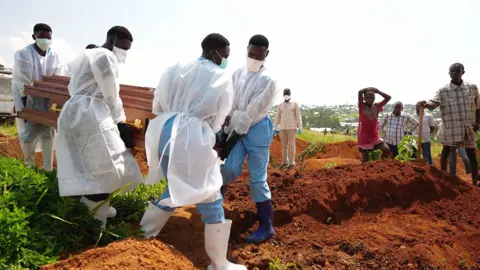 BBC Volunteers in blue scrubs, rubber boots and white gowns carry a coffin towards a dug grave. A few people, including one holding a spade, watch on from the side
