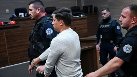 AFP via Getty Images Dušan Maksimović (centre) is seen handcuffed in a courtroom in Pristina after being sentenced to 30 years in prison. Photo: 24 April 2026