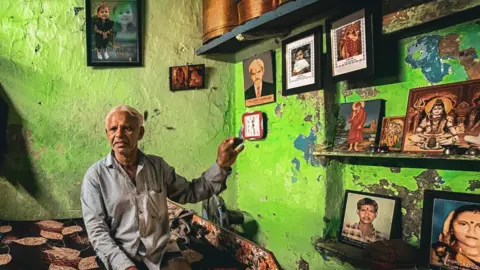 Zoya Mateen Prahlod Thakur sits in his home with framed photographs of his wife, Sarlaben, and granddaughter, Aadhya, who were killed in the Air India crash.