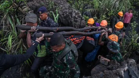 supplied Rescue workers clad in camouflage fatigues and orange helmets carry a body bag balanced on two large poles