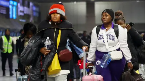 Reuters Two Ghanaian women in winter outfits, one in a Boston sweatshirt, wheeling suitcases at OR Tambo Airport in Johannesburg - 27 May 2026